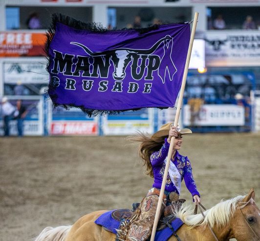 Girl riding a horse in western shirt and cowboy hat while carrying a bright purple Man Up Crusade flag. Community
