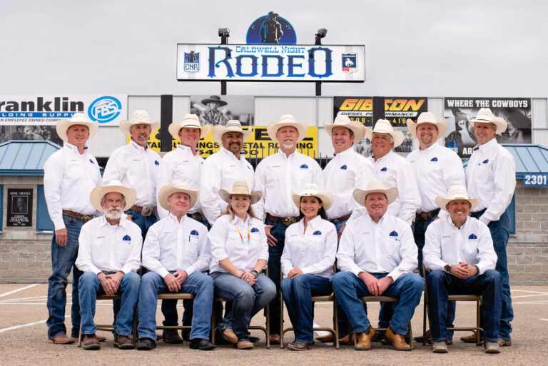 Caldwell Night Rodeo board of directors all in western white shirts with straw cowboy hats and blue jeans. The are standing and seated in front of the front gates to the rodeo grounds with a Caldwell Night Rodeo sign above their heads. Hall of Fame